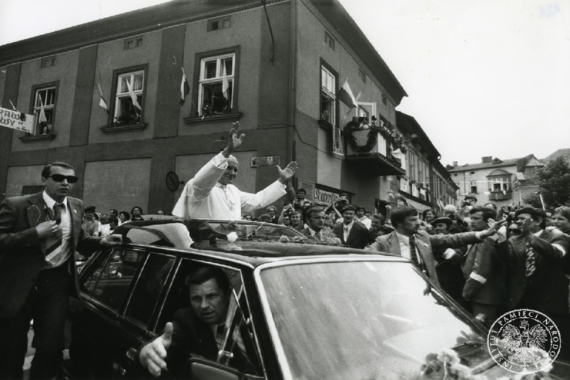 Papież w geście wyciągniętych ramion pozdrawia tłum wiernych przybyłych na wadowicki rynek. Przy samochodzie papieskim idzie służba porządkowa. Na przyległych do rynku budynkach wiszą polskie i watykańskie flagi.