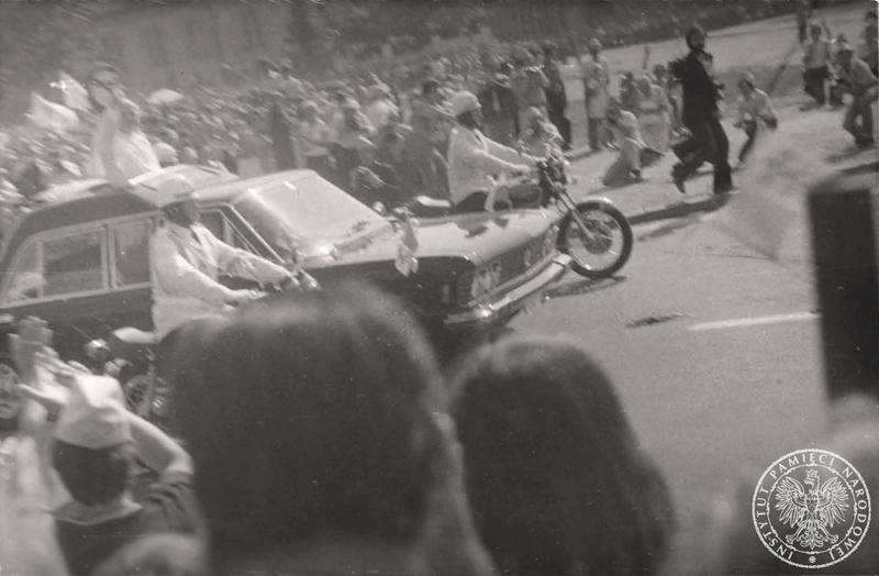 Jan Paweł II pozdrawia zgromadzonych ludzi podczas przejazdu samochodem specjalnym Fiat 130 przez plac Zwycięstwa przed odlotem do Gniezna. W tle budynek na rogu placu Zwycięstwa i ulicy Ossolińskich. 3 VI 1979 r. Sygnatura: IPN Bi 625/39 s. 9
