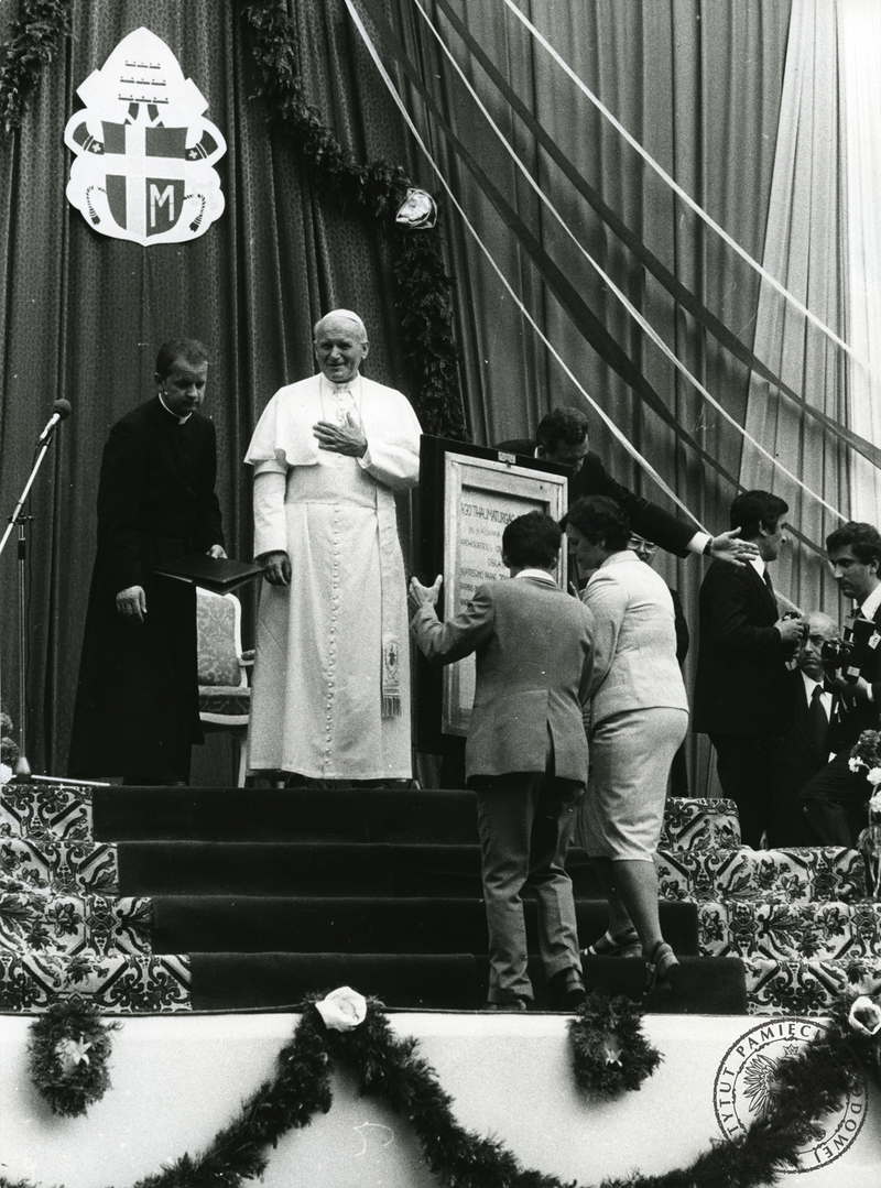 W tle widać płótno ołtarza polowego, na którym wisi herb papieski. Ołtarz polowy przyozdobiony jest w kwiaty i girlandy. Po prawej stronie Papieża stoi ks. Stanisław Dziwisz, a po lewej bp Juliusz Paetz, następnie widać fotoreporterów.