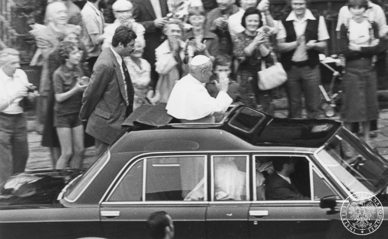 Jan Paweł II pozdrawia wiernych podczas przejazdu samochodem specjalnym Fiat 130 ulicami Krakowa. Zdjęcie wykonane z punktu zakrytego SB na potrzeby dokumentacji operacyjnej. 8 VI 1979 r. Sygnatura: IPNKr-7-7-1-1201
