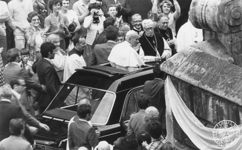 Jan Paweł II w samochodzie specjalnym Fiat 130 podjeżdża pod wejście do Krypty Srebrnych Dzwonów katedry na Wawelu. Wokół samochodu idą pracownicy Biura Ochrony Rządu. Po prawej udekorowany narożnik barokowego muru otaczającego katedrę od południa. 8 VI 1979 r. Sygnatura: IPNKr-7-7-1-1202